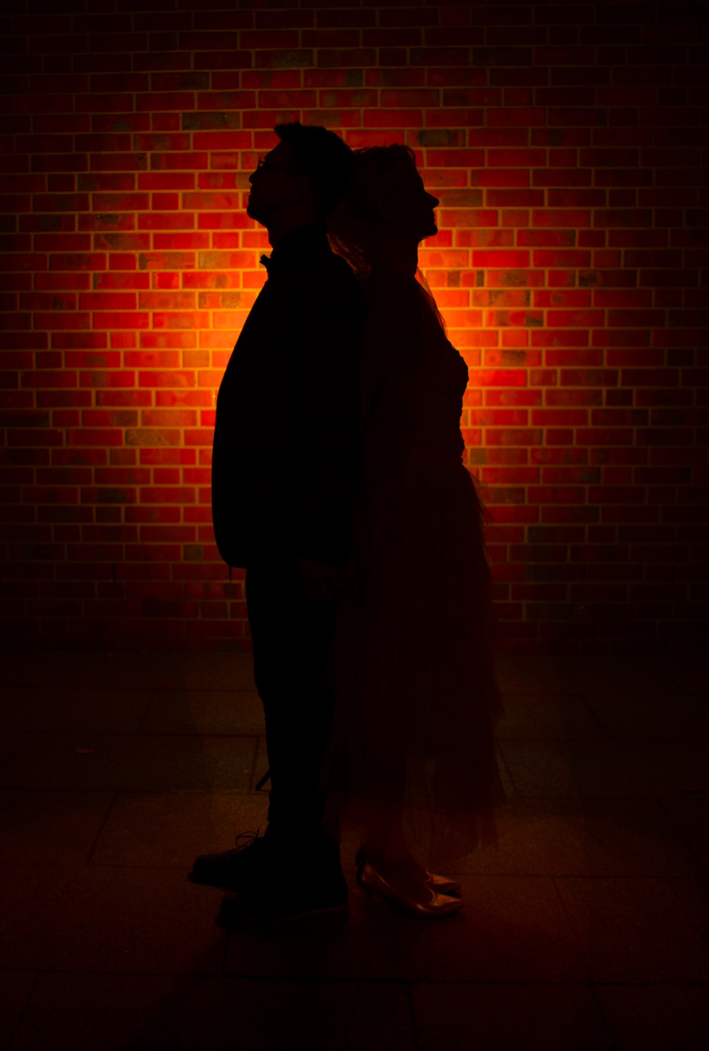 Sylwetki pary na tle ceglanej ściany oświetlonej ciepłym światłem. Mężczyzna w ciemnym ubraniu, kobieta w sukni. Ujęcie w ciemnej tonacji, podkreślające kontury postaci.