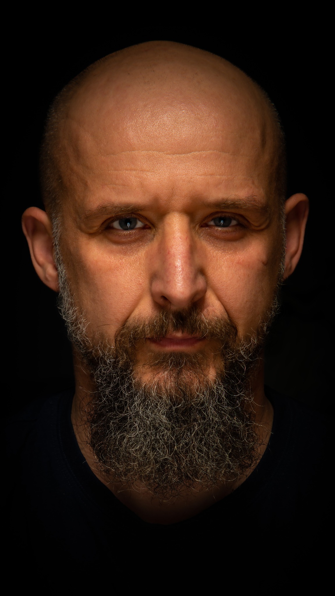 Portret en face mężczyzny z brodą na czarnym tle, oświetlonego z góry. Widoczne zmarszczki i blizna na twarzy, co dodaje charakteru. Ujęcie z bliska, skupiające się na wyrazie twarzy.