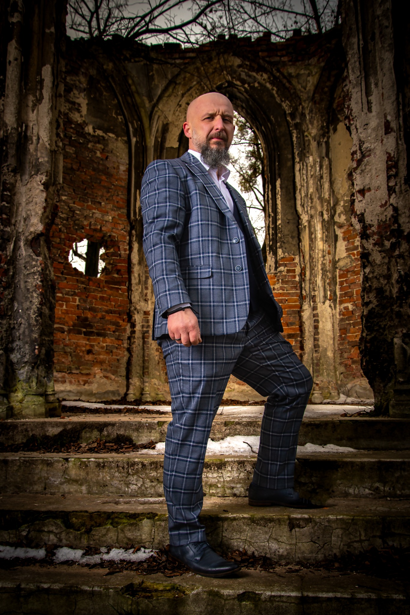 Mężczyzna w garniturze w kratę pozuje na tle ruin z cegły. Ujęcie z dołu podkreśla sylwetkę i architektoniczne tło. Ciemna tonacja i kontrastowe światło.