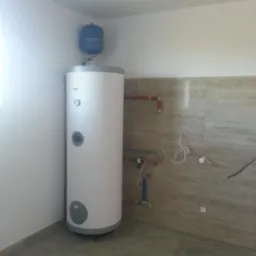 Bojler elektryczny z zbiornikiem wyrównawczym, podłączony do instalacji wodnej w pomieszczeniu z wykończonymi ścianami.