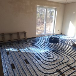 Krzysztof Kubiak - Instalacja ogrzewania podłogowego w jasnym pomieszczeniu z oknem. Widoczne niebieskie rury ułożone na folii z siatką, drabina oparta o ścianę. Etap przed wylaniem posadzki.