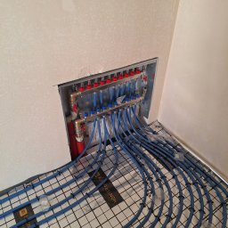 Krzysztof Kubiak - Rozdzielacz ogrzewania podłogowego z niebieskimi rurami ułożonymi na izolacji z rastrem, zamontowany w ścianie w surowym stanie. Widoczne elementy instalacji.