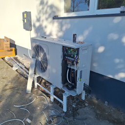 Krzysztof Kubiak - Rozłożona pompa ciepła powietrze-woda na zewnątrz budynku, widoczne elementy instalacji i kable. Na górze puszka z preparatem. Częściowo zdemontowana obudowa.