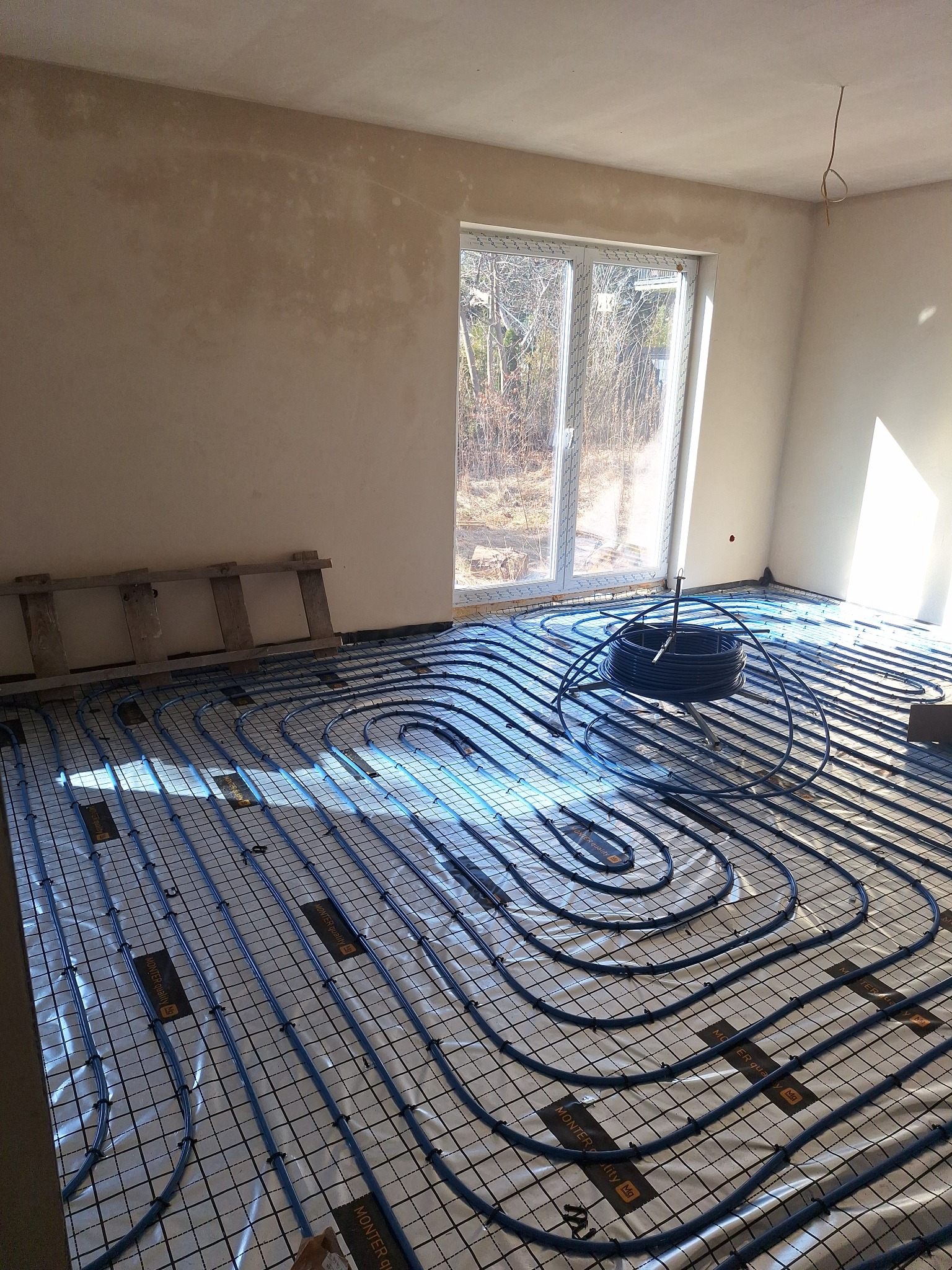 Instalacja ogrzewania podłogowego w jasnym pomieszczeniu z oknem. Widoczne niebieskie rury ułożone na folii z siatką, drabina oparta o ścianę. Etap przed wylaniem posadzki.