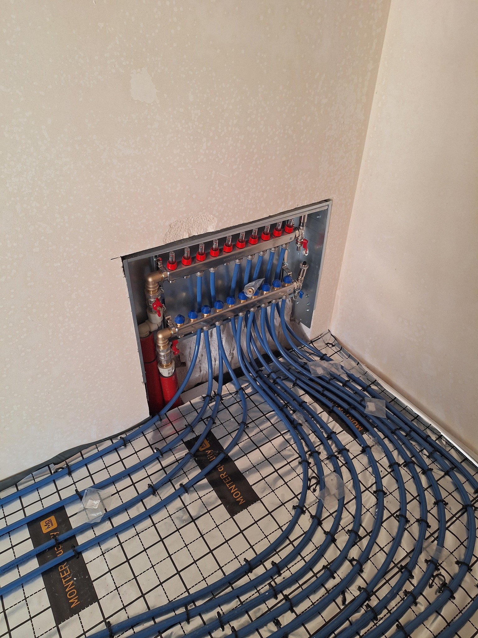 Rozdzielacz ogrzewania podłogowego z niebieskimi rurami ułożonymi na izolacji z rastrem, zamontowany w ścianie w surowym stanie. Widoczne elementy instalacji.