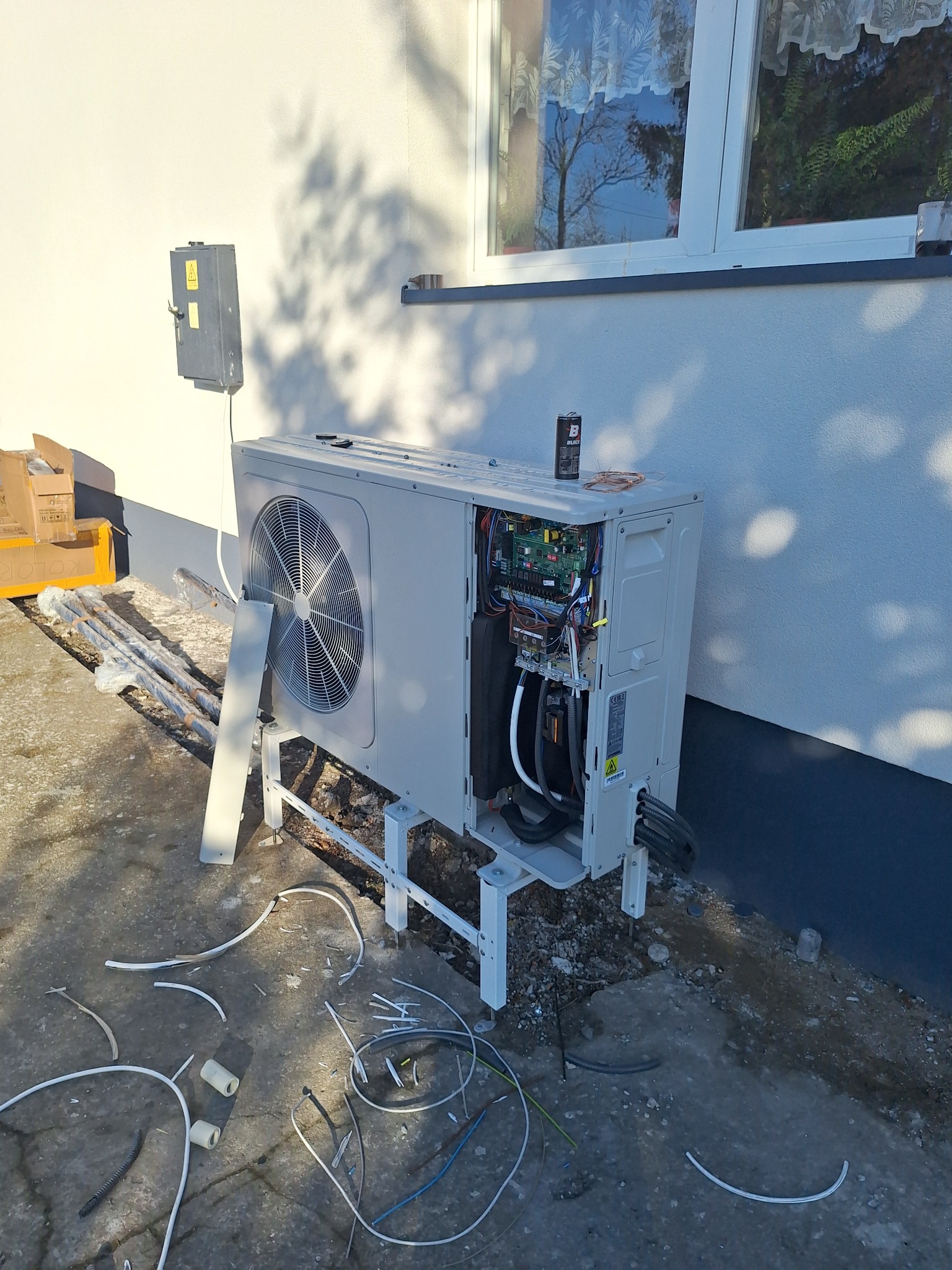 Rozłożona pompa ciepła powietrze-woda na zewnątrz budynku, widoczne elementy instalacji i kable. Na górze puszka z preparatem. Częściowo zdemontowana obudowa.