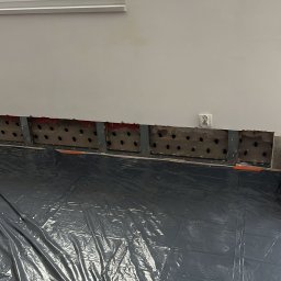 POR INVEST PLUS SPÓŁKA Z OGRANICZONĄ ODPOWIEDZIALNOŚCIĄ - Przygotowanie do izolacji ściany: odsłonięta konstrukcja z otworami, zabezpieczona folią podłoga, widoczne gniazdko elektryczne. Prace remontowe w pomieszczeniu.