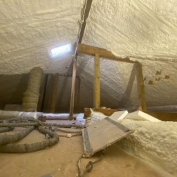 POR INVEST PLUS SPÓŁKA Z OGRANICZONĄ ODPOWIEDZIALNOŚCIĄ - Poddasze po ociepleniu pianką PUR, widoczne drewniane elementy konstrukcyjne i instalacje. Biała, nierówna powierzchnia pianki pokrywa skośne ściany i sufit.