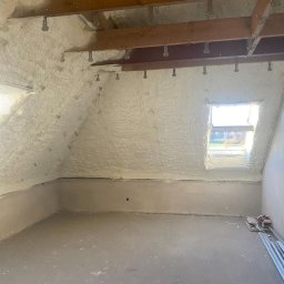 POR INVEST PLUS SPÓŁKA Z OGRANICZONĄ ODPOWIEDZIALNOŚCIĄ - Poddasze po ociepleniu pianką PUR, widoczne drewniane belki stropowe z metalowymi mocowaniami i okno dachowe. Ściany i sufit pokryte pianą, podłoga surowa.