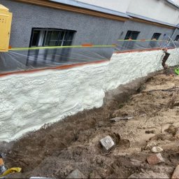 POR INVEST PLUS SPÓŁKA Z OGRANICZONĄ ODPOWIEDZIALNOŚCIĄ - Izolacja fundamentów pianką PUR, widoczne zabezpieczenie folią i taśmą. Żółta skrzynka gazowa na ścianie budynku. Wykop wzdłuż fundamentu.