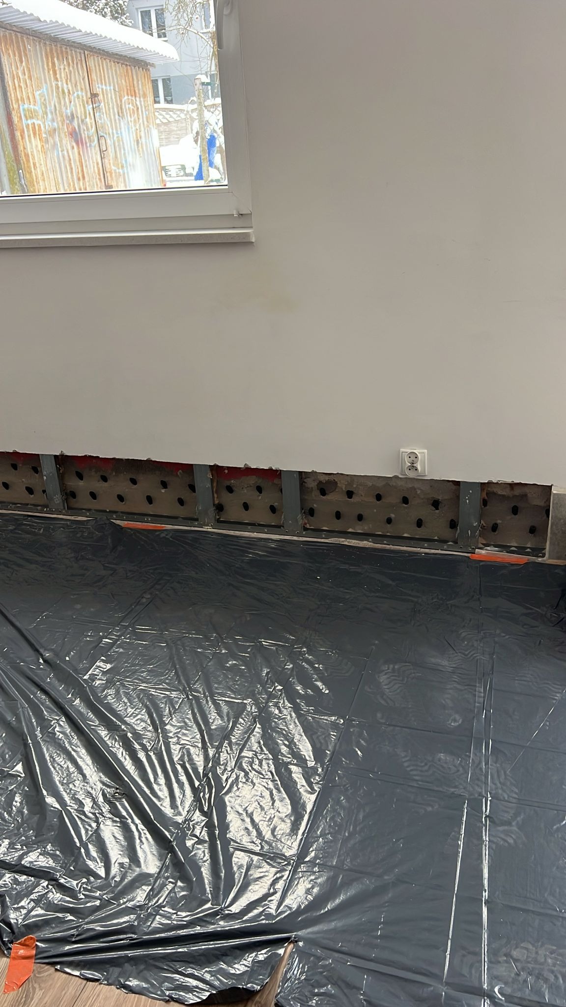 Przygotowanie do izolacji ściany: odsłonięta konstrukcja z otworami, zabezpieczona folią podłoga, widoczne gniazdko elektryczne. Prace remontowe w pomieszczeniu.