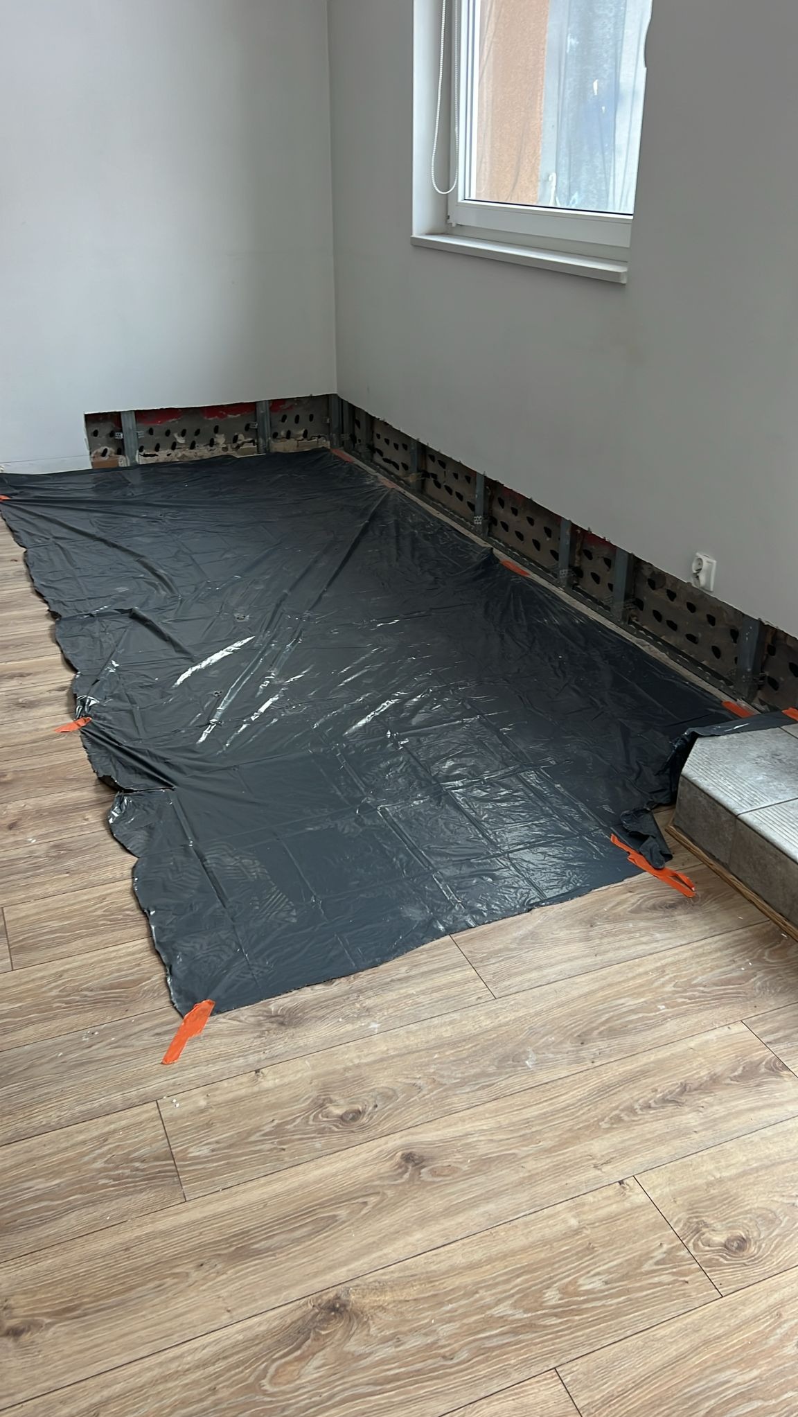 Przygotowanie do prac wykończeniowych: zabezpieczona podłoga folią malarską, odsłonięta konstrukcja ściany z widocznymi profilami i fragmentami instalacji. Jasne wnętrze.