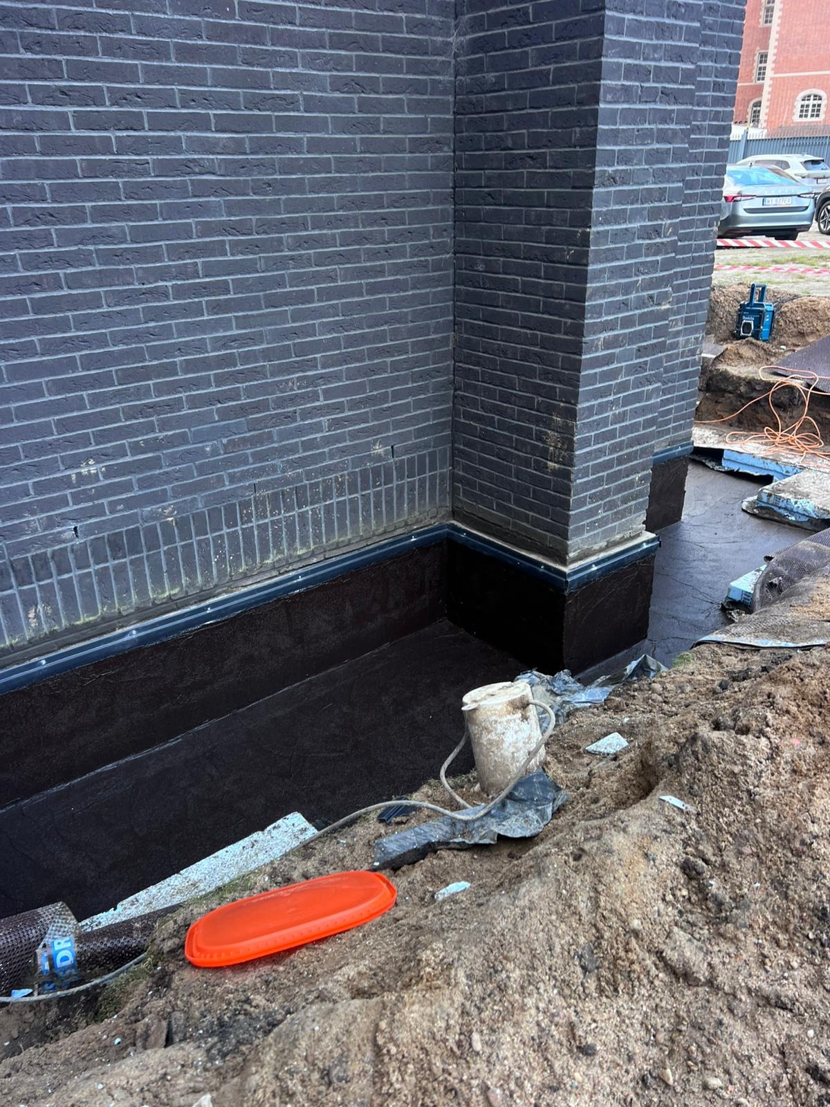 Izolacja pionowa fundamentu budynku z ciemnej cegły, zabezpieczona czarną powłoką hydroizolacyjną. Widoczne narzędzia i materiały budowlane w wykopie.