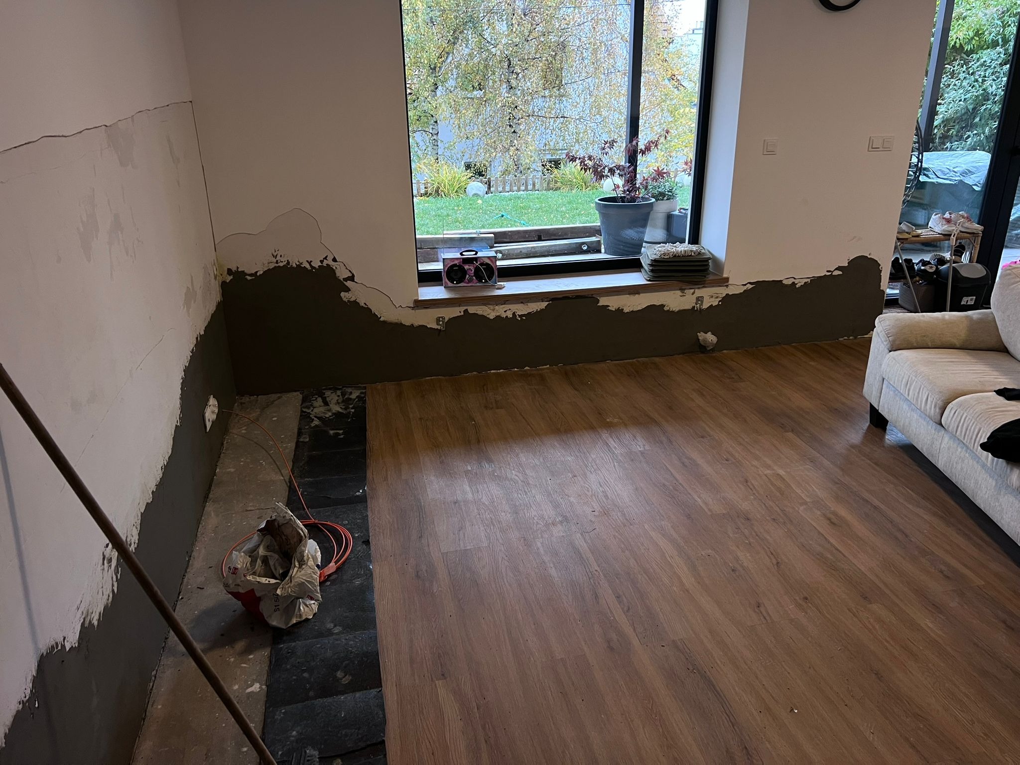 Przygotowanie ścian do malowania w pomieszczeniu z oknem i drewnianą podłogą. Widoczne ślady szpachlowania przy podłodze i częściowo na ścianie. Część podłogi jeszcze nie wykończona.