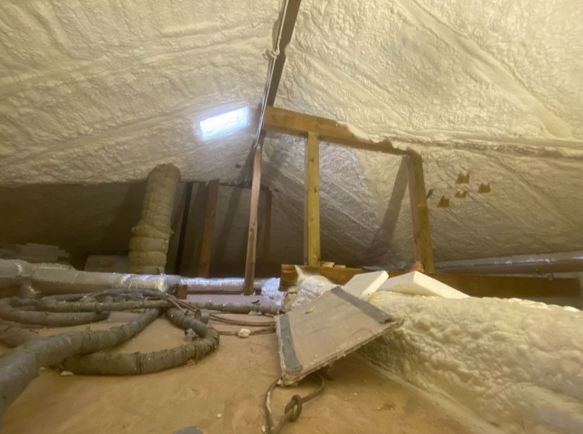 Poddasze po ociepleniu pianką PUR, widoczne drewniane elementy konstrukcyjne i instalacje. Biała, nierówna powierzchnia pianki pokrywa skośne ściany i sufit.