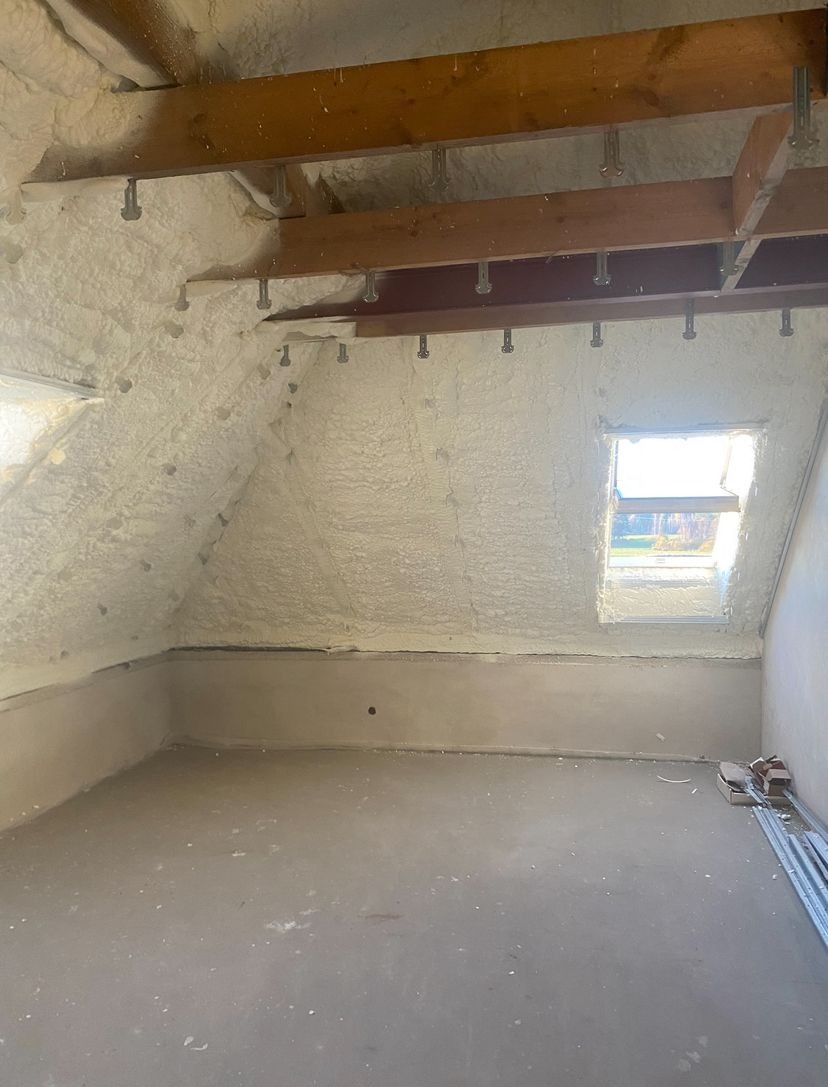 Poddasze po ociepleniu pianką PUR, widoczne drewniane belki stropowe z metalowymi mocowaniami i okno dachowe. Ściany i sufit pokryte pianą, podłoga surowa.