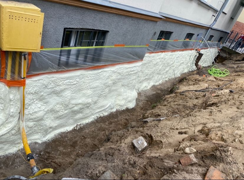 Izolacja fundamentów pianką PUR, widoczne zabezpieczenie folią i taśmą. Żółta skrzynka gazowa na ścianie budynku. Wykop wzdłuż fundamentu.