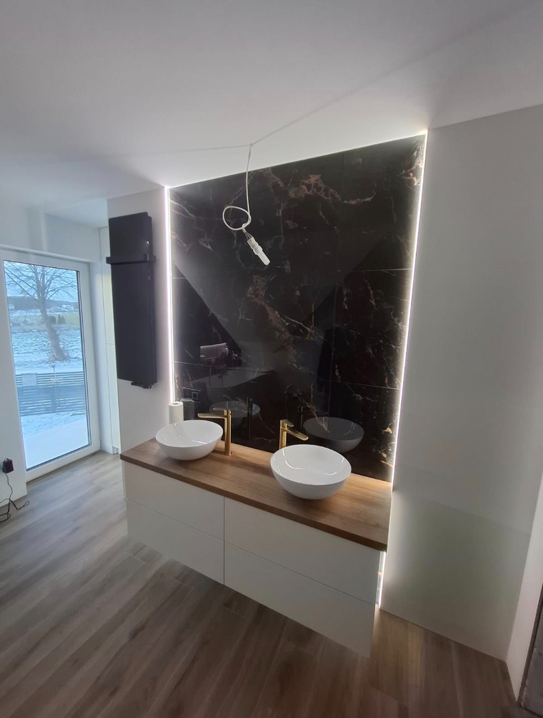 Nowoczesna łazienka z podwójną umywalką nablatową, drewnianym blatem i czarną ścianą z marmurowym wzorem, podświetloną listwą LED. Widok na okno.