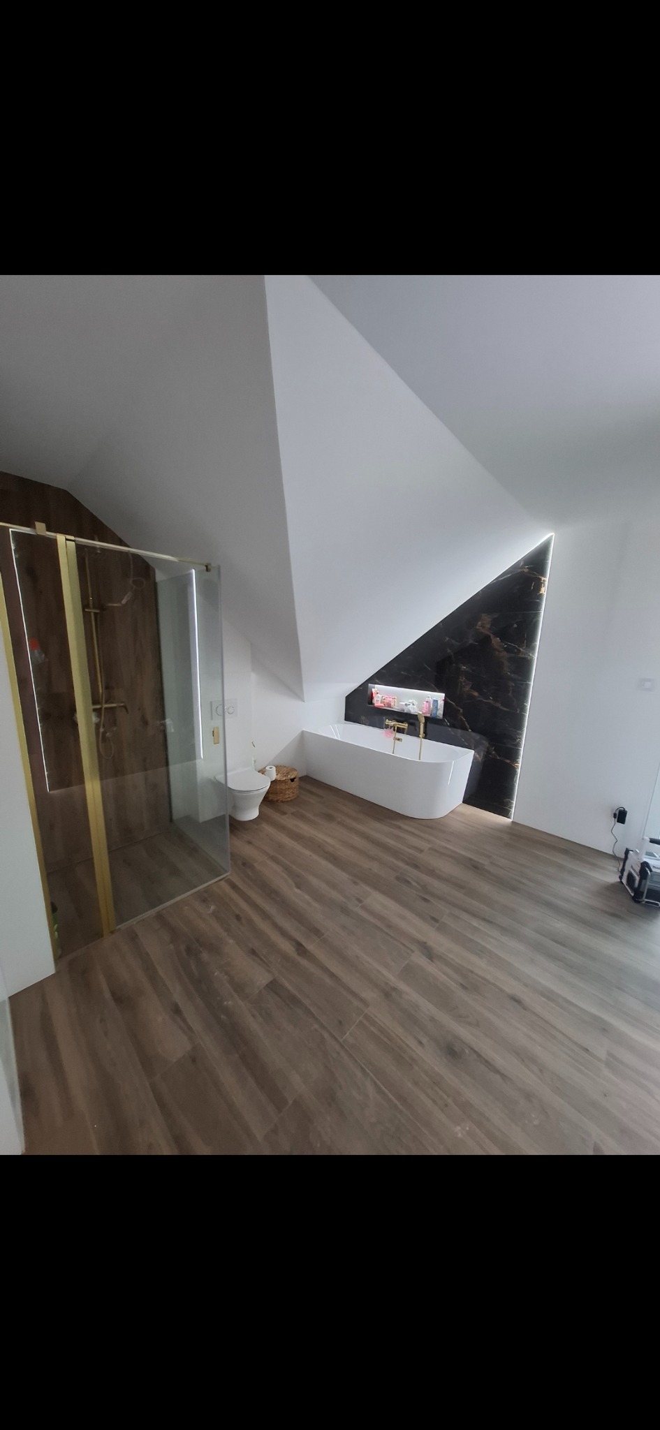 Nowoczesna łazienka na poddaszu z kabiną prysznicową ze złotymi detalami, wolnostojącą wanną i czarnym marmurem podświetlonym LED. Drewnopodobna podłoga.