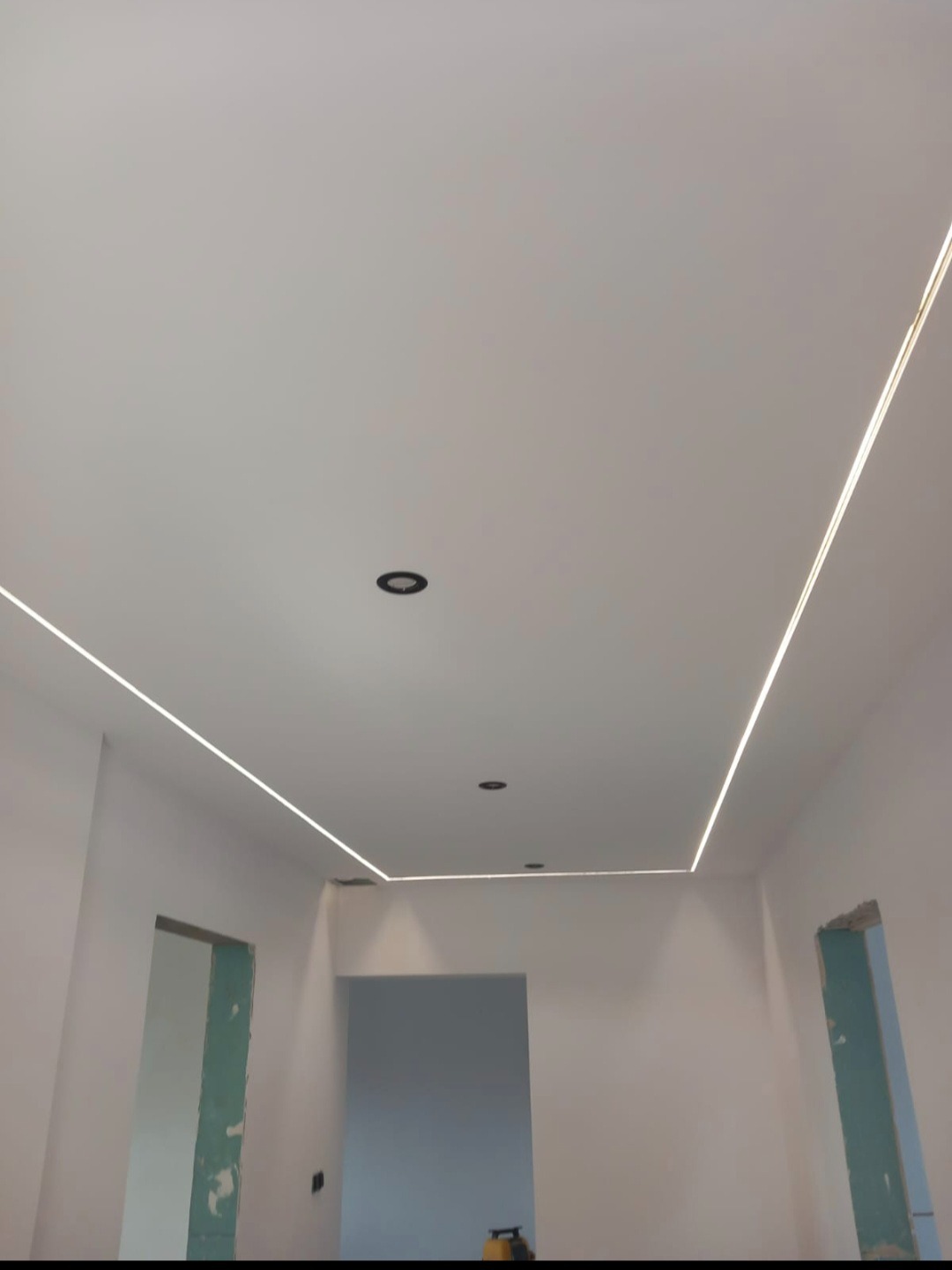 Białe wnętrze z podwieszanym sufitem i liniowym oświetleniem LED. Widoczne otwory drzwiowe i sufitowe lampy punktowe. Minimalistyczny design.