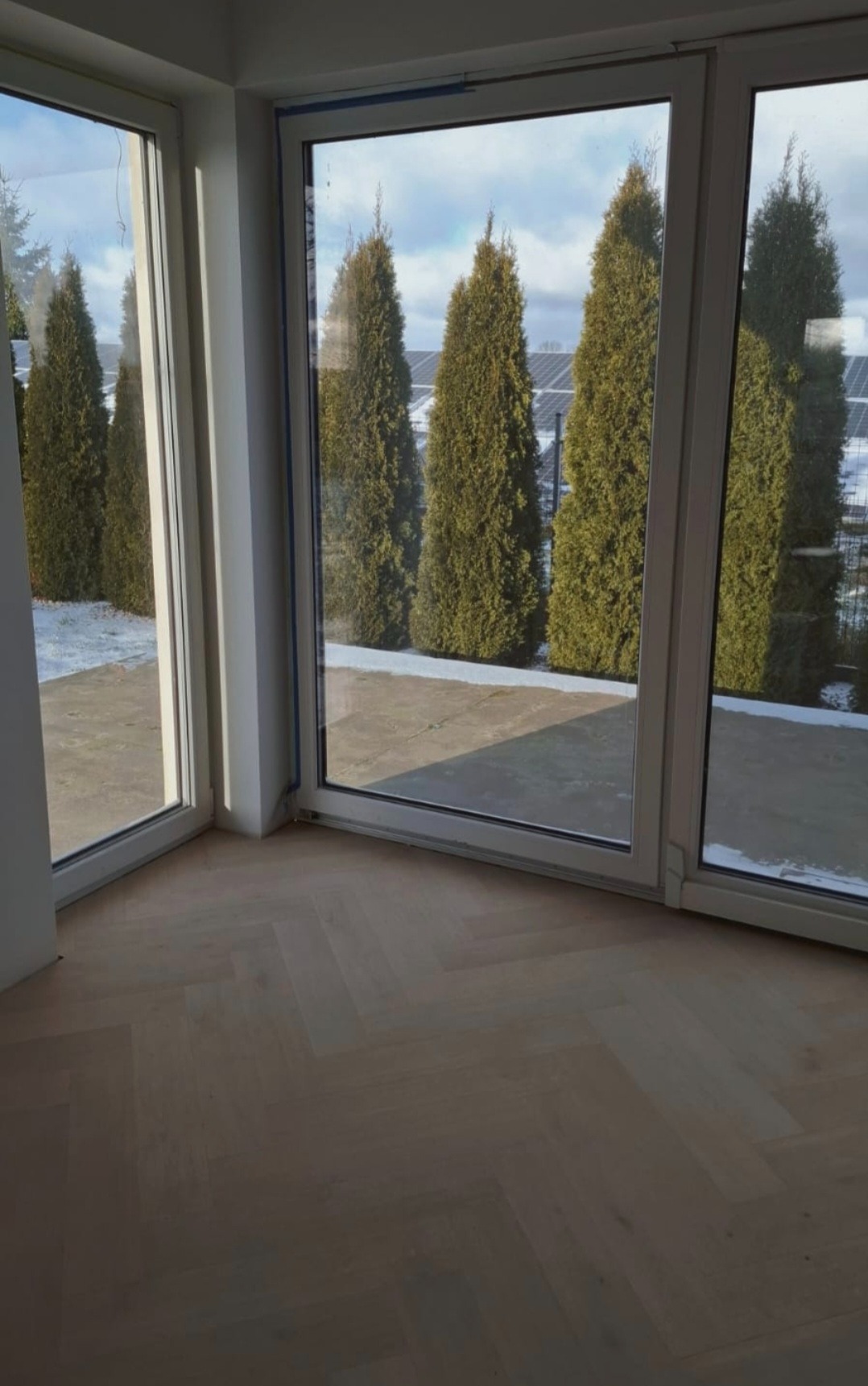 Jasny parkiet ułożony w jodełkę francuską w nowym wnętrzu z dużymi oknami tarasowymi, przez które widać rząd tuj i panele słoneczne w tle.