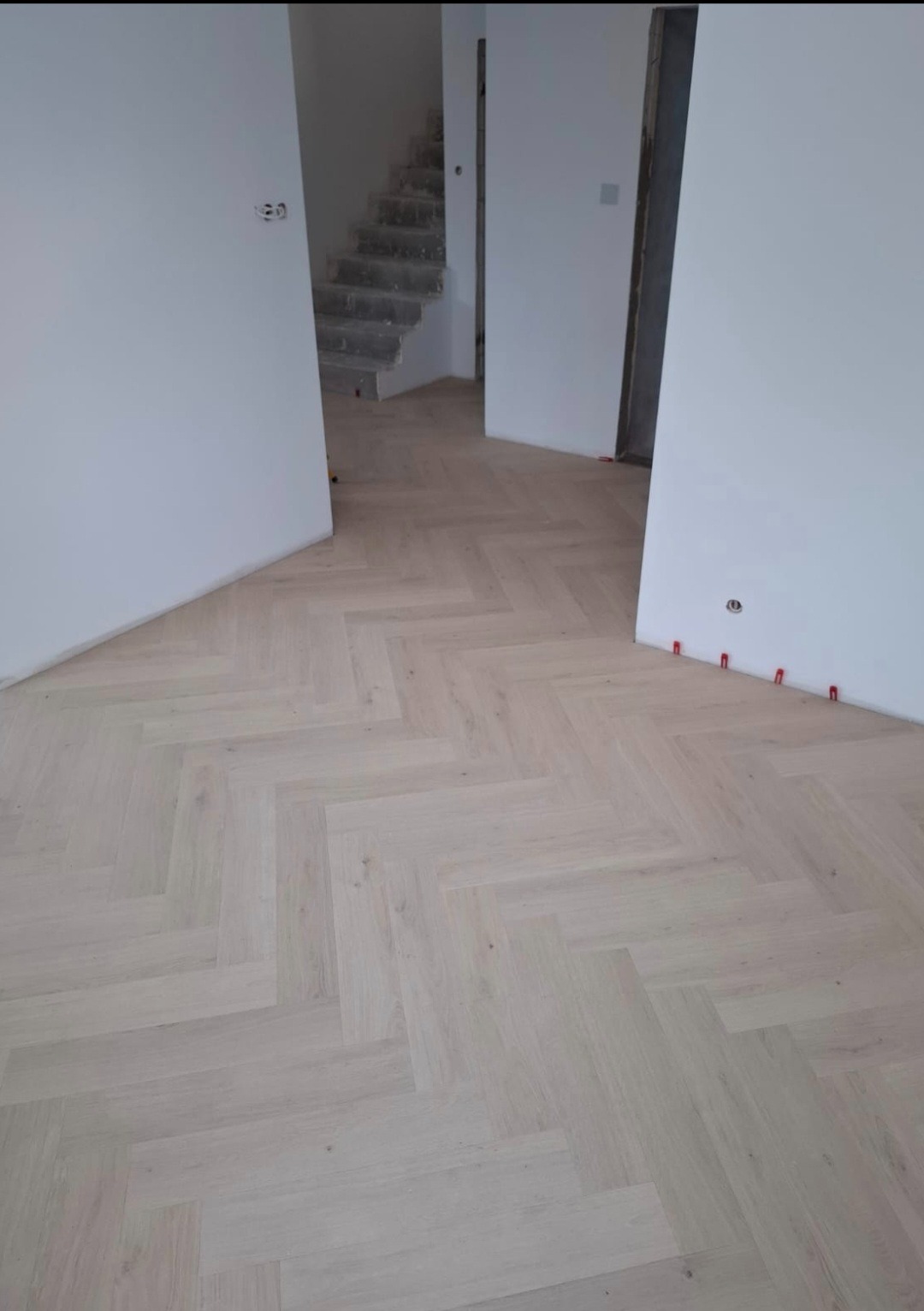 Jasne panele ułożone w jodełkę francuską, widoczne czerwone kliny dystansowe przy ścianie, surowe ściany i schody w tle. Minimalistyczne wnętrze w trakcie wykańczania.