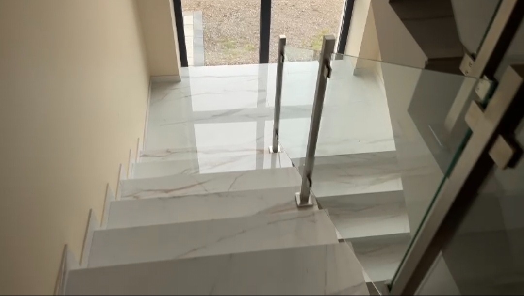 Białe marmurowe schody z metalową balustradą i szklanym wypełnieniem, widok z góry, jasne wnętrze z oknem w tle. Nowoczesny design.