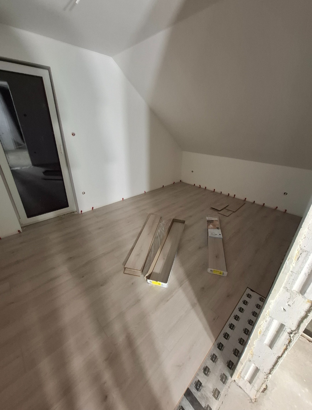 Układanie paneli podłogowych w pokoju na poddaszu. Widoczne panele, listwy przypodłogowe i elementy montażowe. Ściany białe, skos dachu.