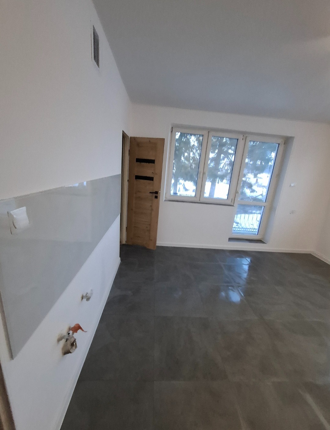 Nowoczesne wnętrze po remoncie: szara podłoga, białe ściany z instalacją, drewniane drzwi i okno z widokiem na drzewa. Minimalistyczny design.