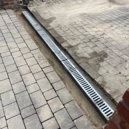 MagicMan - Ułożona kostka brukowa z zamontowanym odpływem liniowym, widoczne piaskowe podsypki. Częściowo ułożona nawierzchnia, ceglany murek w tle.