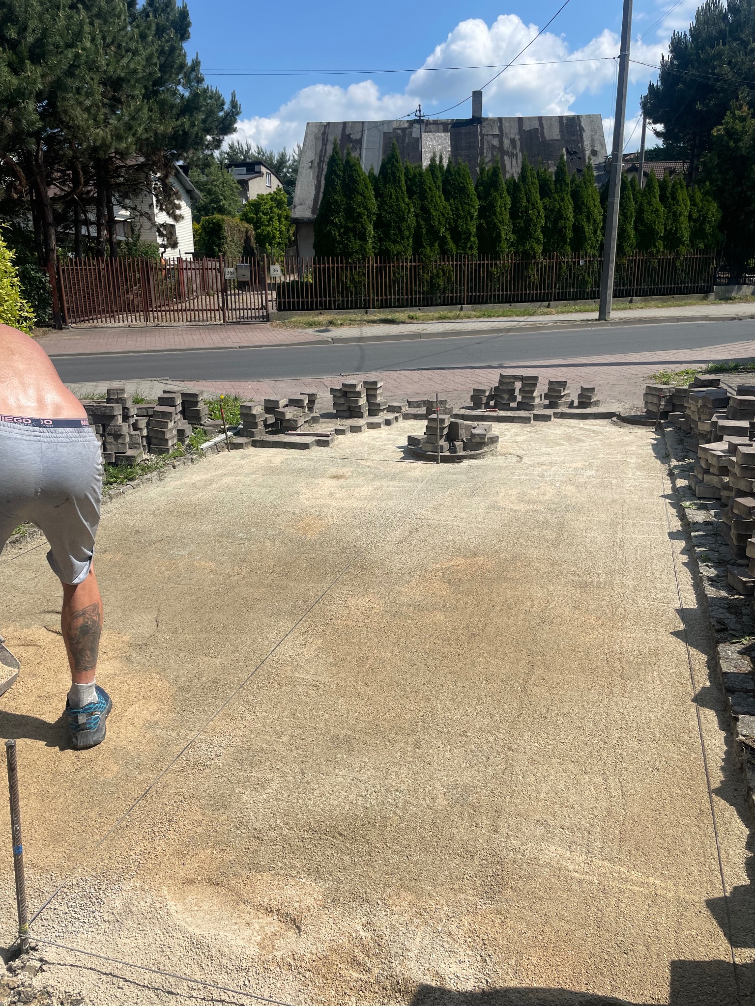 Przygotowanie podłoża pod kostkę brukową: wyrównany teren z piaskiem, ułożone krawężniki i stosy kostki. Widoczny fragment sylwetki brukarza z tatuażem na łydce.