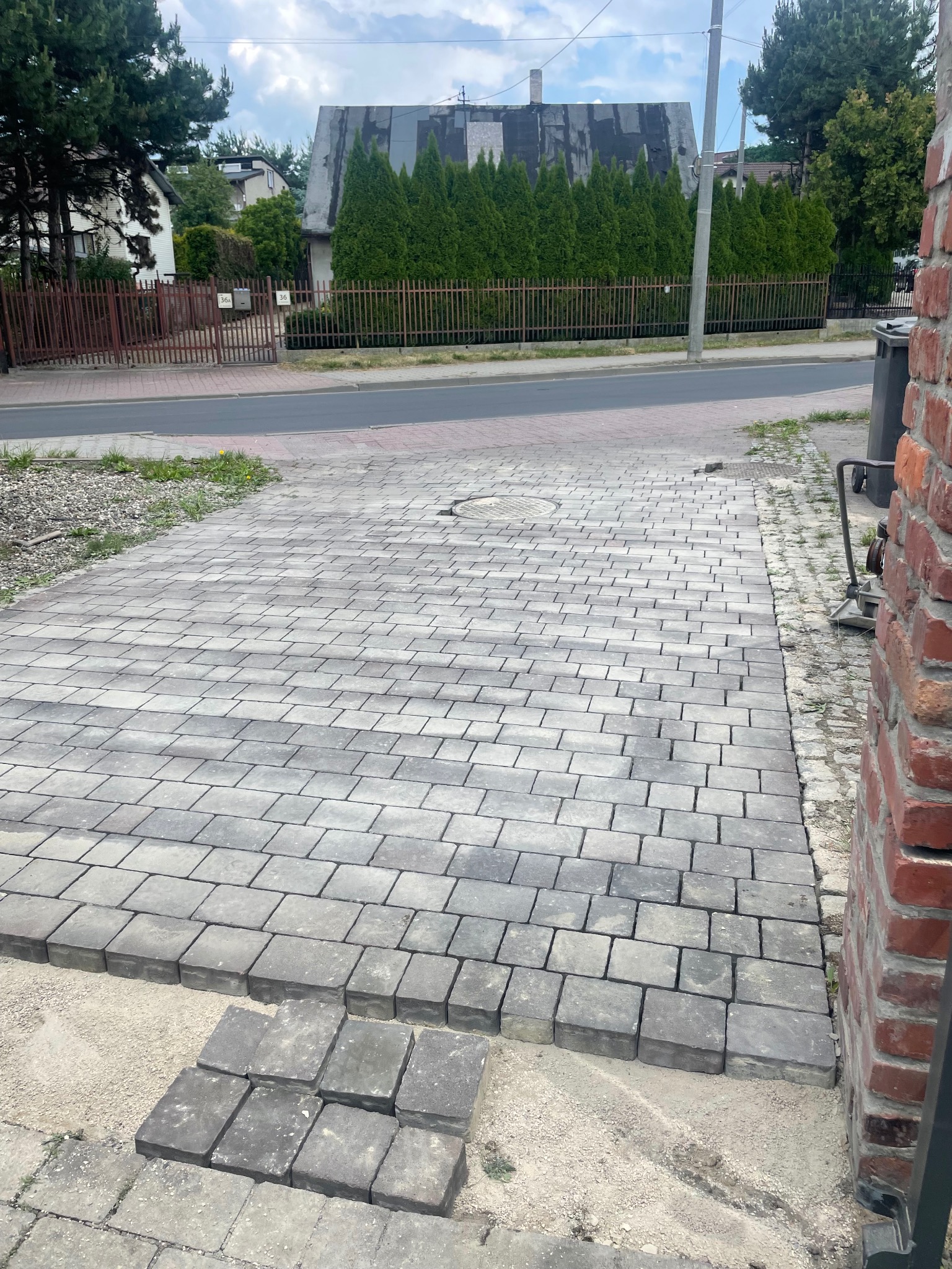 Ułożona szara kostka brukowa na podjeździe, widoczne niedokończone fragmenty i przygotowane kostki. W tle dom z zielonymi tujami i metalowe ogrodzenie.