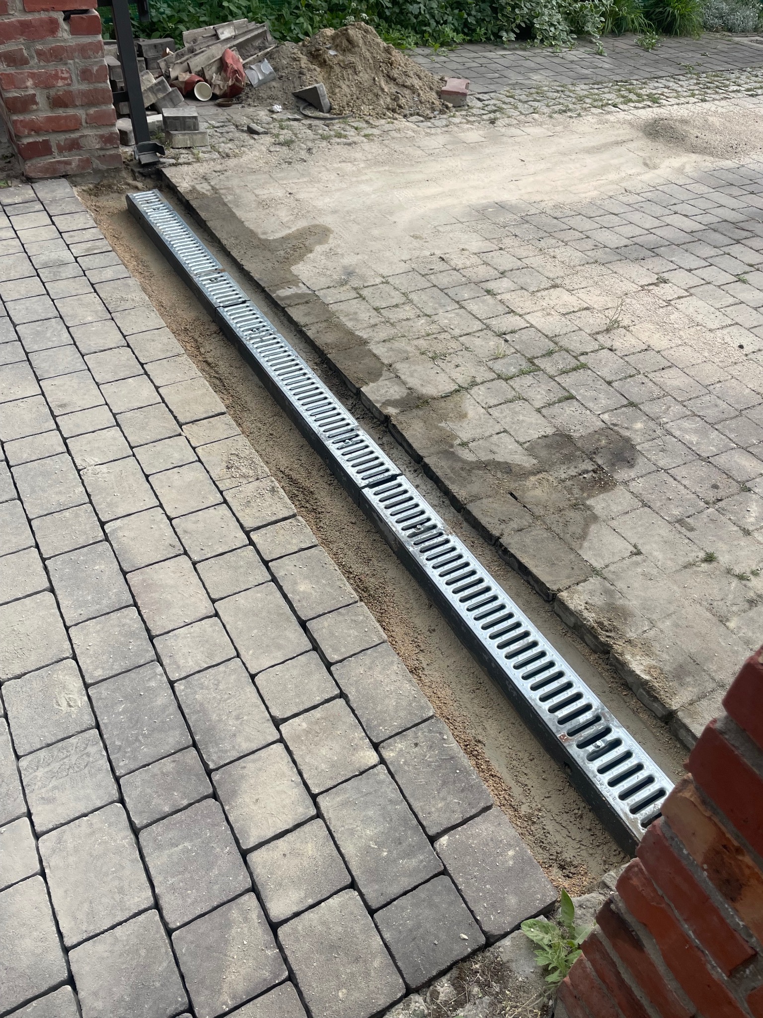 Ułożona kostka brukowa z zamontowanym odpływem liniowym, widoczne piaskowe podsypki. Częściowo ułożona nawierzchnia, ceglany murek w tle.