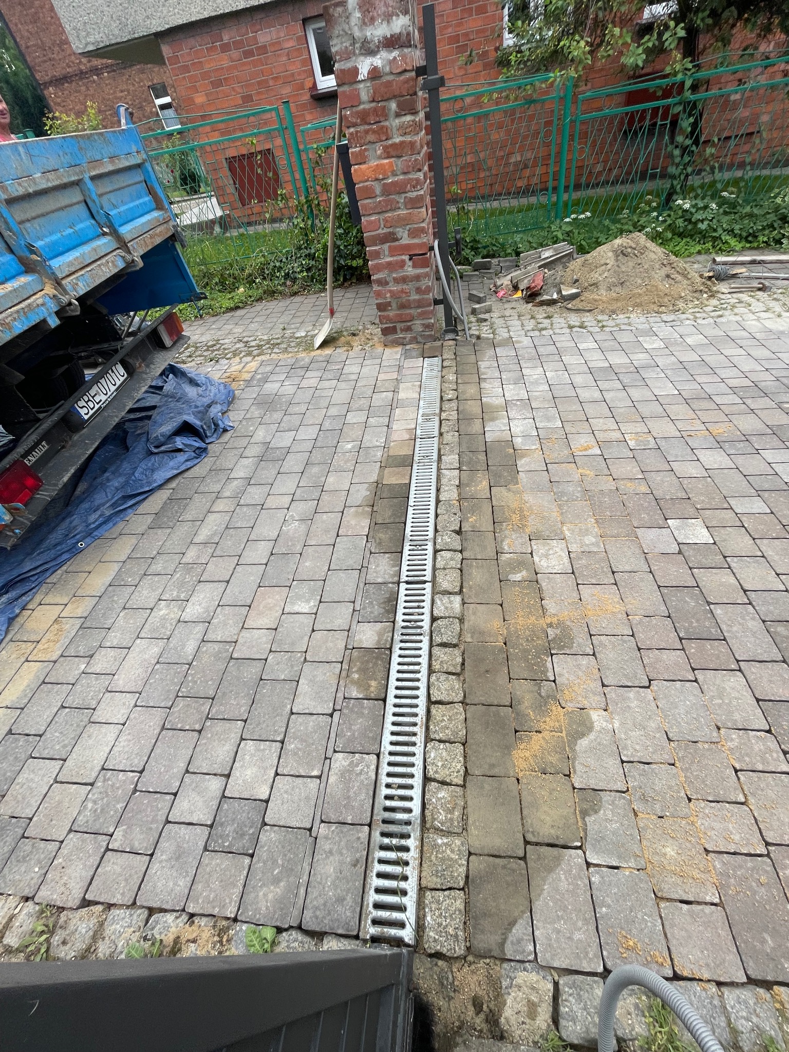 Ułożona kostka brukowa z odpływem liniowym na posesji. Widoczny fragment ciężarówki i narzędzia. Ceglany słupek ogrodzeniowy w tle.