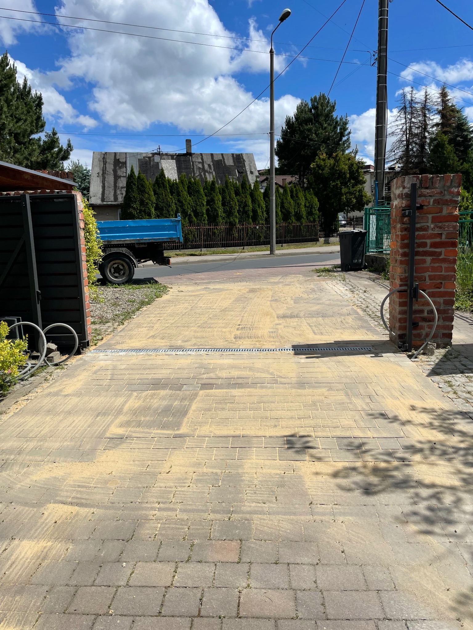 Ułożona kostka brukowa z widocznym odpływem liniowym, brama wjazdowa z ceglanym słupkiem i zaparkowana przyczepa w tle.