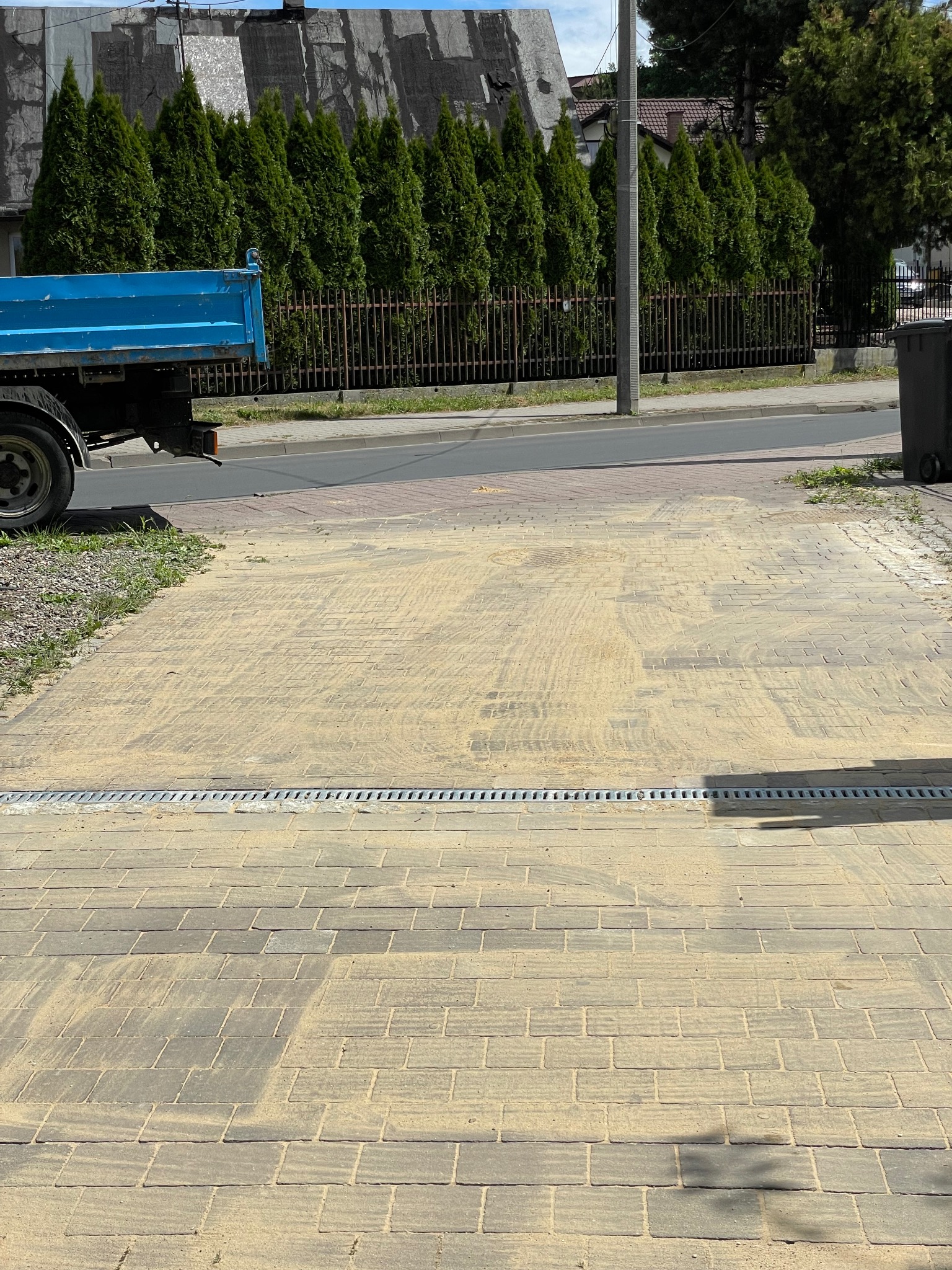 Ułożona kostka brukowa z odpływem liniowym, zasypana piaskiem. W tle widoczny samochód ciężarowy i rząd tuj wzdłuż ogrodzenia.