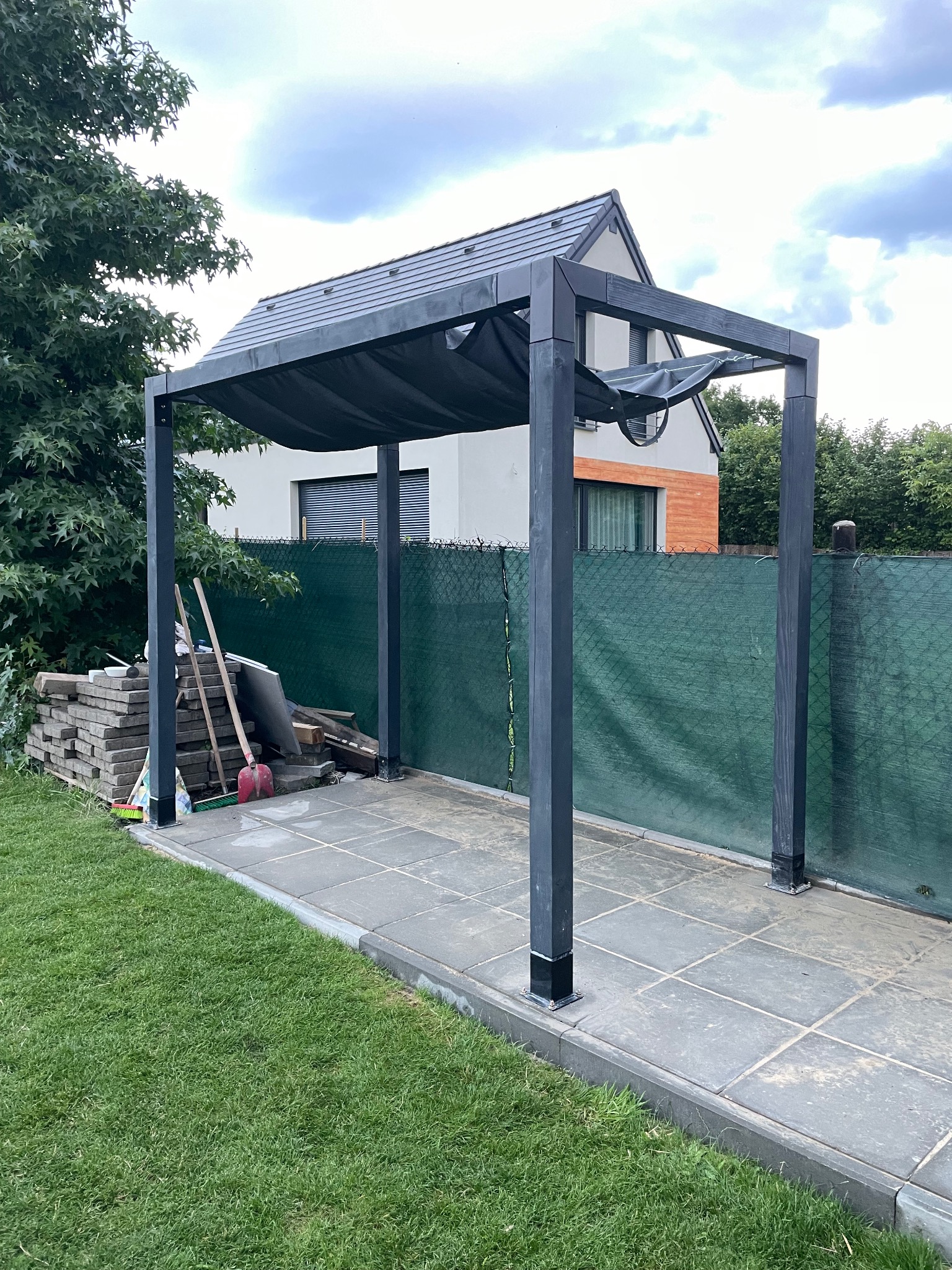 Nowoczesna, czarna pergola z zadaszeniem na tle zielonego ogrodzenia i domu. Widoczne ułożone płyty chodnikowe i narzędzia budowlane obok konstrukcji.