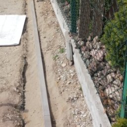 Zbyszek - Wykopany rów z betonowymi obrzeżami wzdłuż siatki ogrodzeniowej, przygotowany pod ścieżkę lub podjazd. Widoczny plan na ziemi i kamienie w tle.