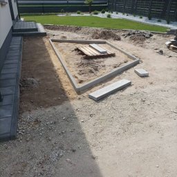 Zbyszek - Przygotowanie podłoża pod kostkę brukową: obrzeża ułożone w kwadrat, widoczne płyty na palecie, trawnik w tle. Etap prac ziemnych przed ułożeniem nawierzchni.