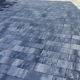 Zbyszek - Ułożona kostka brukowa w odcieniach szarości, z widocznym fragmentem ogrodzenia panelowego w tle. Na pierwszym planie stos kostki brukowej. Widok z góry.