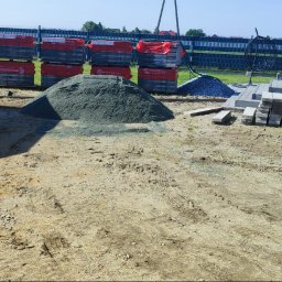 Zbyszek - Przygotowany teren pod kostkę brukową: stosy kostki, piasek, żwir. W tle widoczny fragment tarasu i ogrodzenie panelowe.