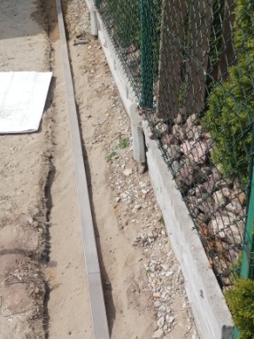 Wykopany rów z betonowymi obrzeżami wzdłuż siatki ogrodzeniowej, przygotowany pod ścieżkę lub podjazd. Widoczny plan na ziemi i kamienie w tle.