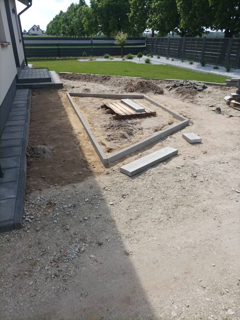 Przygotowanie podłoża pod kostkę brukową: obrzeża ułożone w kwadrat, widoczne płyty na palecie, trawnik w tle. Etap prac ziemnych przed ułożeniem nawierzchni.