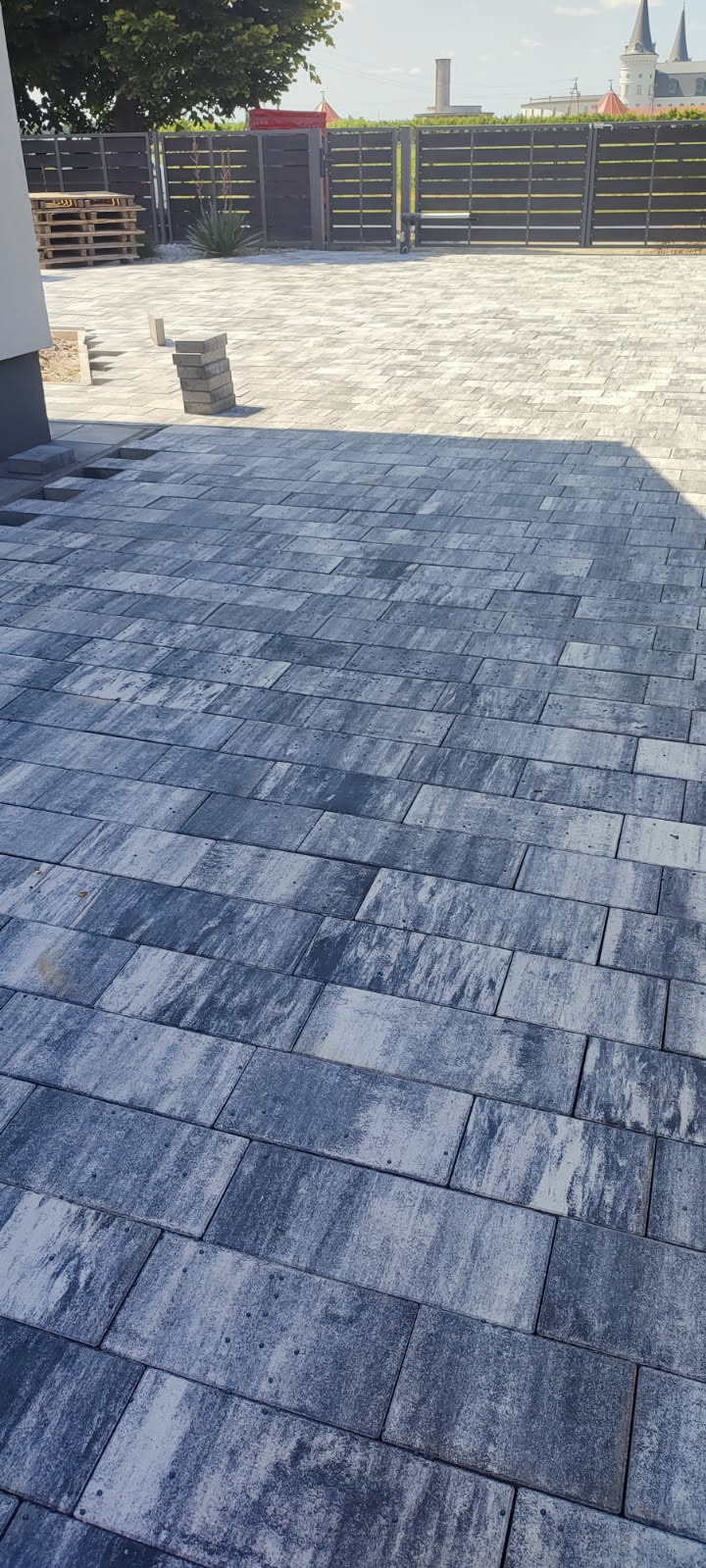Ułożona kostka brukowa w odcieniach szarości, z widocznym fragmentem ogrodzenia panelowego w tle. Na pierwszym planie stos kostki brukowej. Widok z góry.