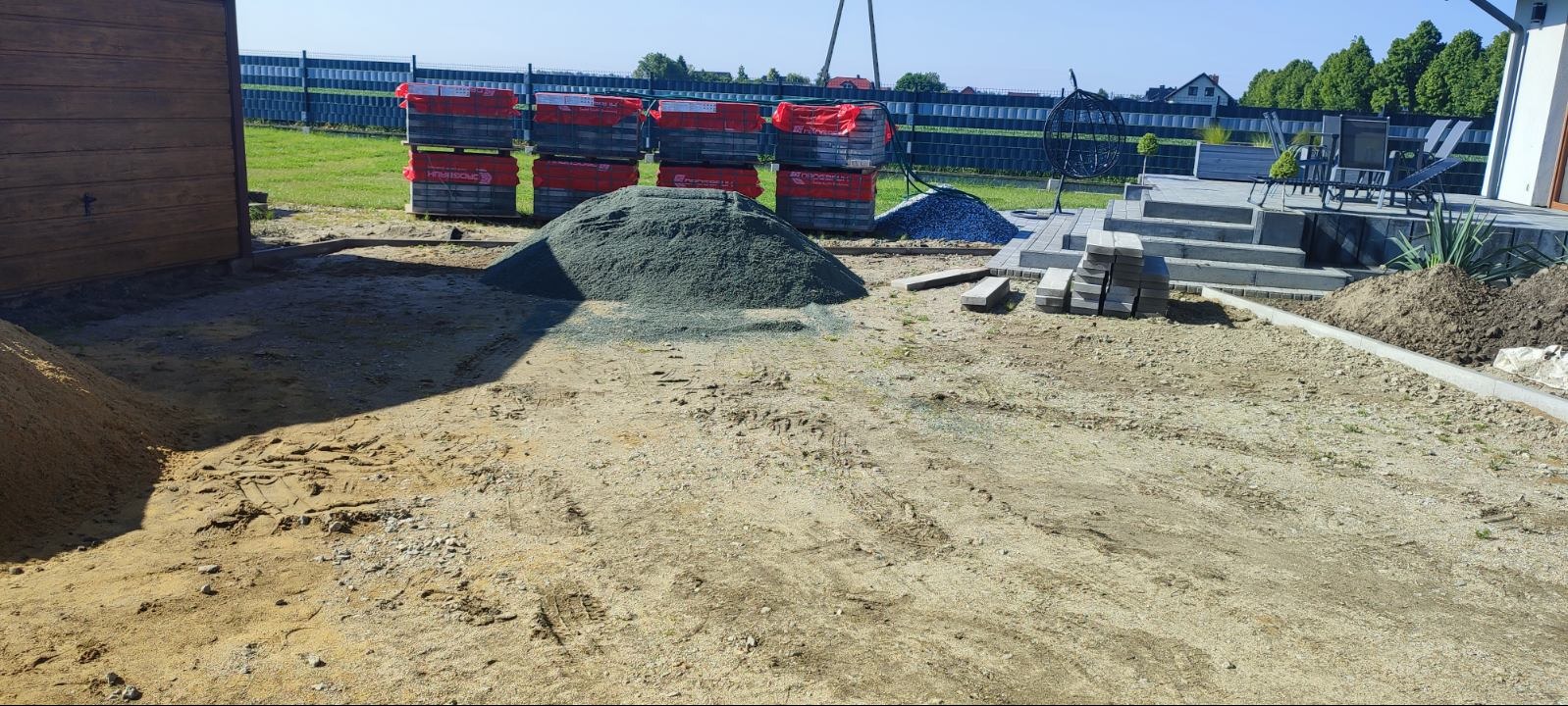 Przygotowany teren pod kostkę brukową: stosy kostki, piasek, żwir. W tle widoczny fragment tarasu i ogrodzenie panelowe.