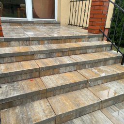 Dominik Milejski Transport & Construction - Schody wejściowe wyłożone kostką brukową w odcieniach szarości i beżu, prowadzące do drzwi z oknem. Widoczna ceglana kolumna i fragment metalowej balustrady.