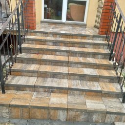 Dominik Milejski Transport & Construction - Schody wejściowe wyłożone kostką brukową w odcieniach szarości i beżu, z czarną metalową balustradą i ceglanymi słupkami. Widok z dołu, trawa u podstawy schodów.
