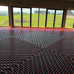 REM-BUD INSTAL BŁAŻEJ PĘDZIMĄŻ, ERYK PĘDZIMĄŻ SPÓŁKA CYWILNA - Czerwone rury ogrzewania podłogowego ułożone na czarnej macie w surowym wnętrzu z widokiem na zielone wzgórza przez duże okna.