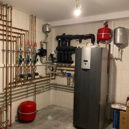 REM-BUD INSTAL BŁAŻEJ PĘDZIMĄŻ, ERYK PĘDZIMĄŻ SPÓŁKA CYWILNA - Złożona instalacja CO w kotłowni: nowoczesny piec, liczne miedziane rury, zawory, manometry i zbiorniki wyrównawcze. Białe płytki na ścianach i szara podłoga.