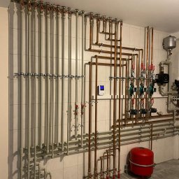 REM-BUD INSTAL BŁAŻEJ PĘDZIMĄŻ, ERYK PĘDZIMĄŻ SPÓŁKA CYWILNA - Złożona instalacja grzewcza z rur miedzianych i szarych, zaworów oraz czerwonego zbiornika wyrównawczego na tle białych płytek. Widoczny manometr i sterownik.